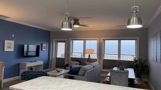 A modern living room with a kitchen area, large windows with a view of the ocean, and a ceiling fan. - The Surf - steps to the beach (Hampton)