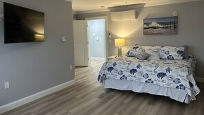 A bedroom with a bed, a ceiling fan, a TV, a painting, and a door leading to another room. - The Surf - steps to the beach (Hampton)