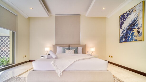 A large bed with a headboard, two bedside tables with lamps, a large abstract painting on the wall, and a view of greenery outside.