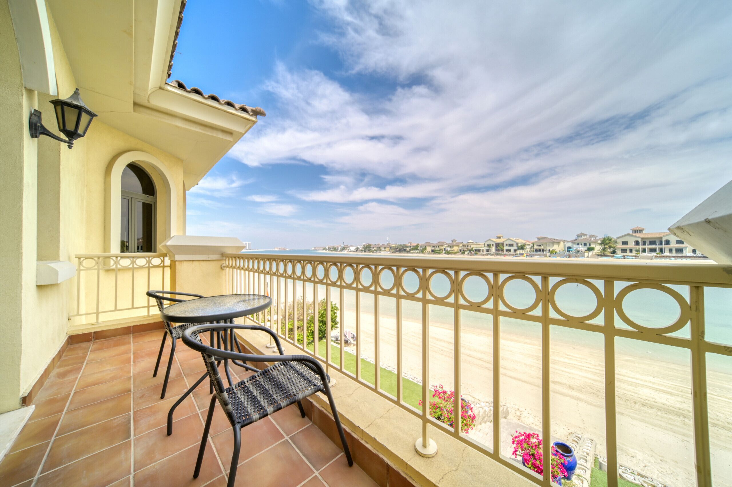 A balcony with a table and chairs, a railing, and a view of the beach and distant buildings.
