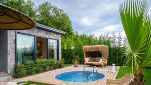 Un área de piscina moderna al aire libre con sillas de descanso, una zona de estar con techo de paja y un edificio de piedra con puertas de cristal.