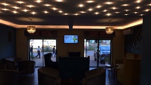 A hotel lobby with a large screen displaying a sports event, multiple seating areas, and a ceiling with numerous lights. - LIEBE HOTEL (Yaoundé)