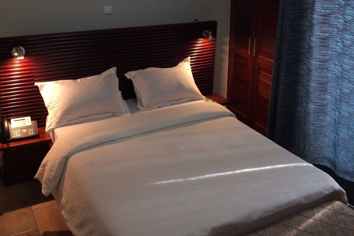Una cama bien tendida con sábanas blancas, cabecera de madera, mesita de noche con lámpara y un cuadro en la pared.