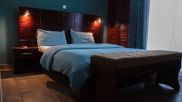 A neatly made bed with a headboard, a bedside table with a lamp, and a bench at the foot of the bed. - LIEBE HOTEL (Yaoundé)
