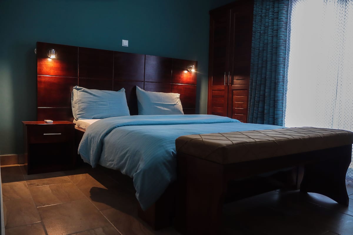 Una cama bien hecha con cabecera, una mesita de noche con lámpara y un banco al pie de la cama.