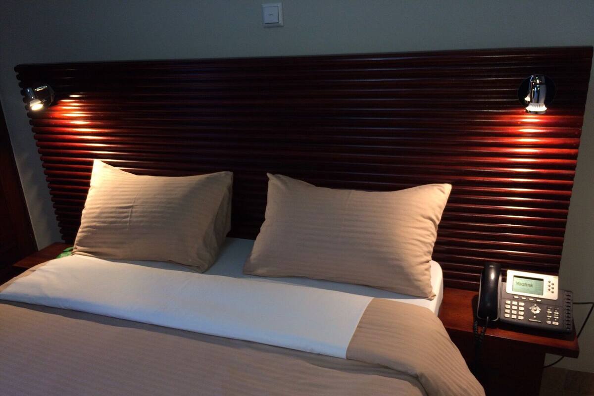 Habitación de hotel con cabecera de madera, dos almohadas color beige, una colcha blanca y un teléfono en la mesita de noche.