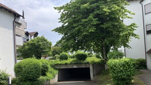 Een verhard pad dat leidt naar een overdekte ingang, omgeven door weelderige begroeiing en woongebouwen.