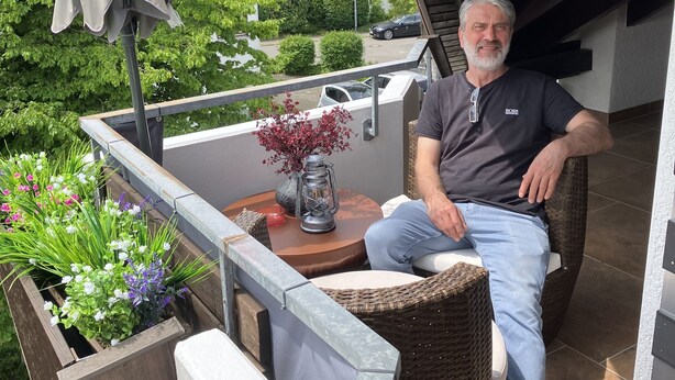 Een man zit op een rieten stoel op een balkon met uitzicht op een tuin en een huis.