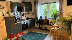 Een kleine keuken met moderne apparatuur, een eethoek met een houten tafel en stoel, en uitzicht op groen buiten.