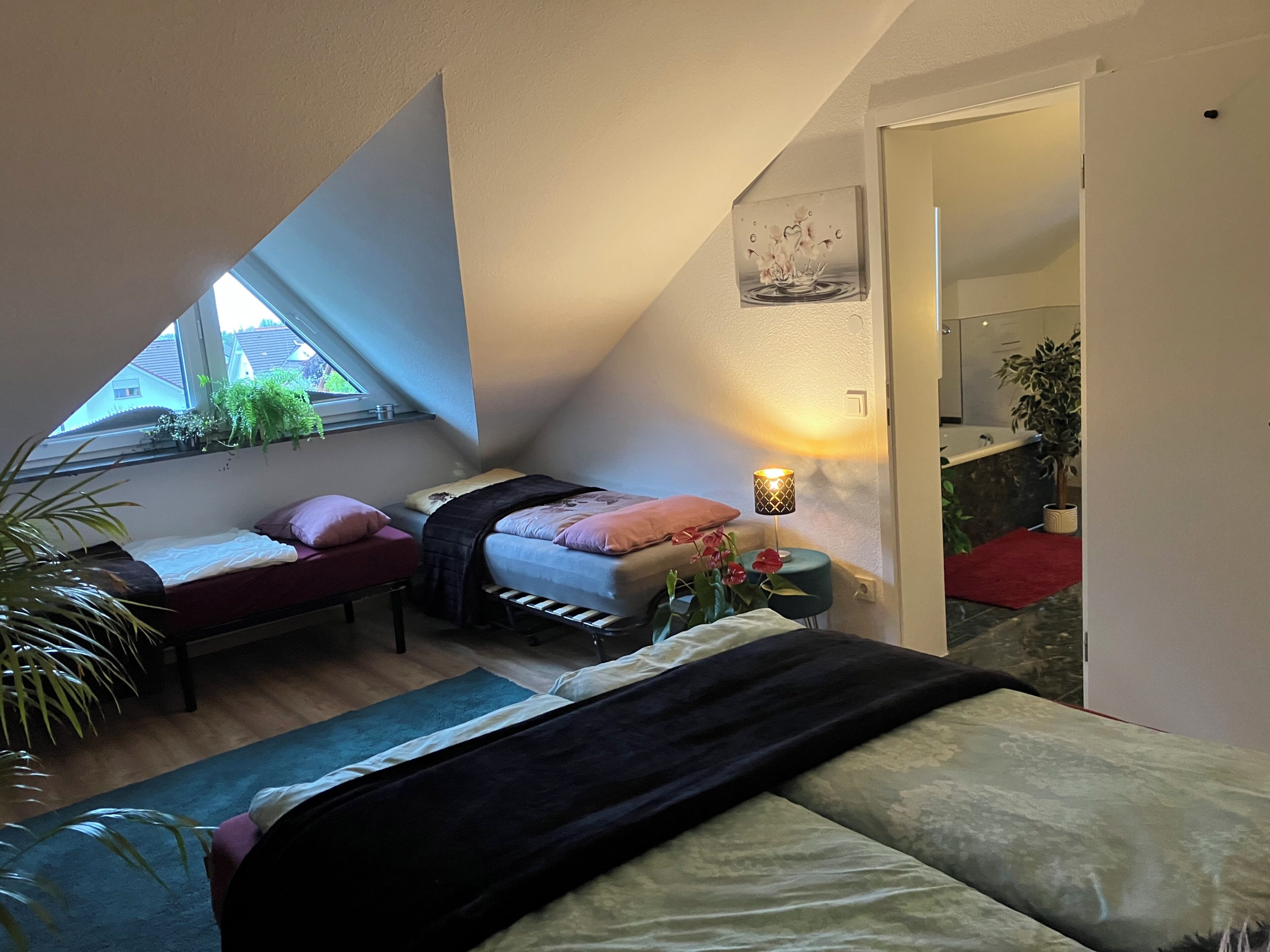 A bedroom with a bed, bedside table, lamp, and a window with plants.