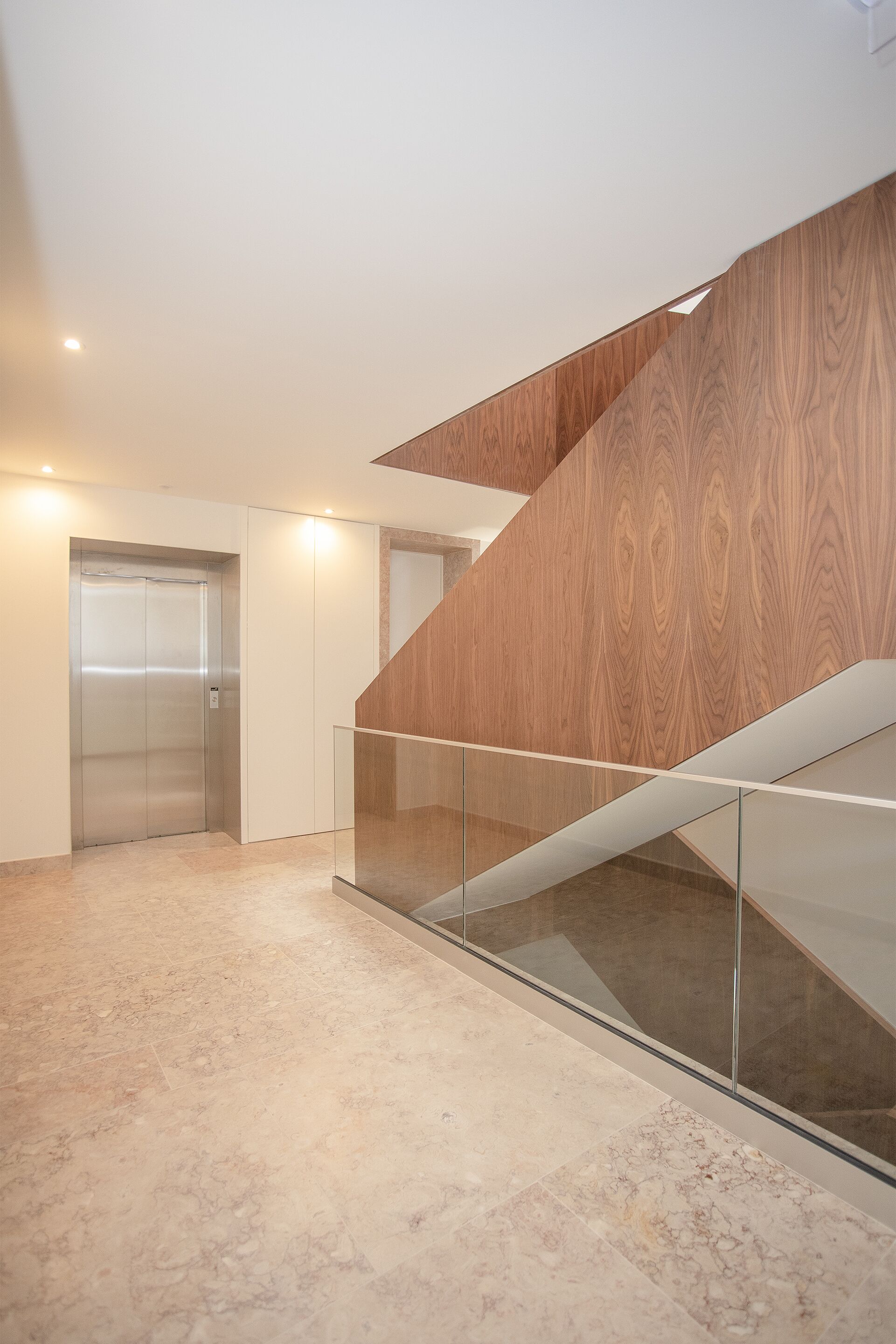A modern interior with a glass staircase, elevators, and a marble floor.