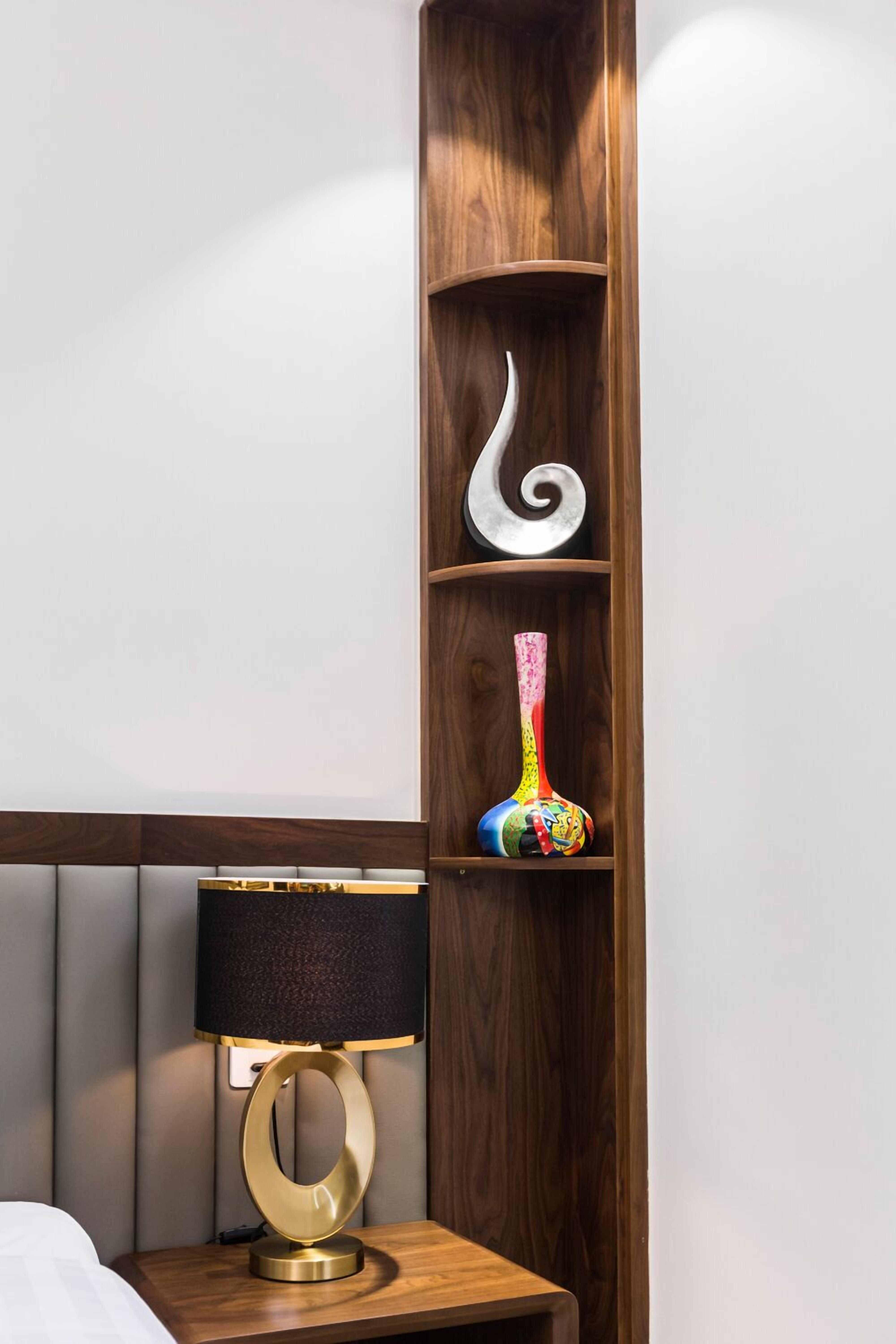 Ein Nachttisch aus Holz mit einer modernen Lampe, einer Dekovase und einer weißen Spiralplastik auf einem Regal.