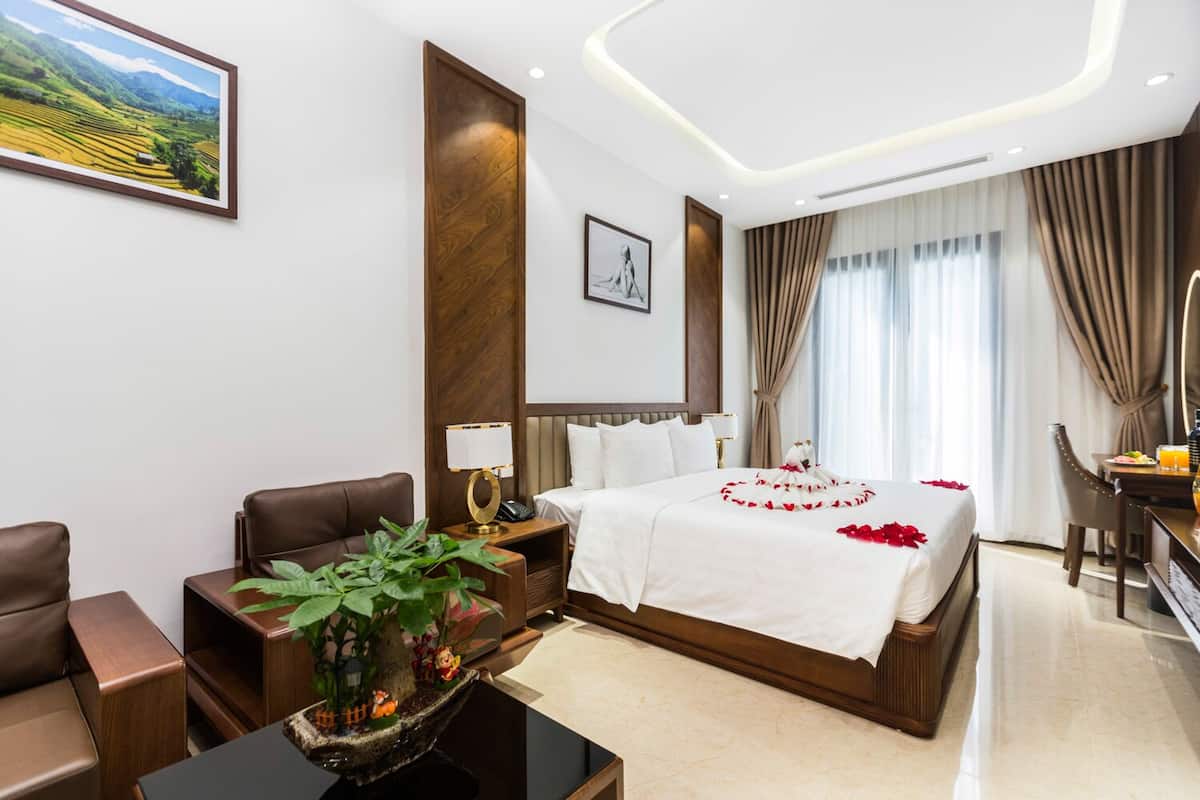 A hotel room with a large bed, a desk, a chair, a small table with a plant, and a painting on the wall.