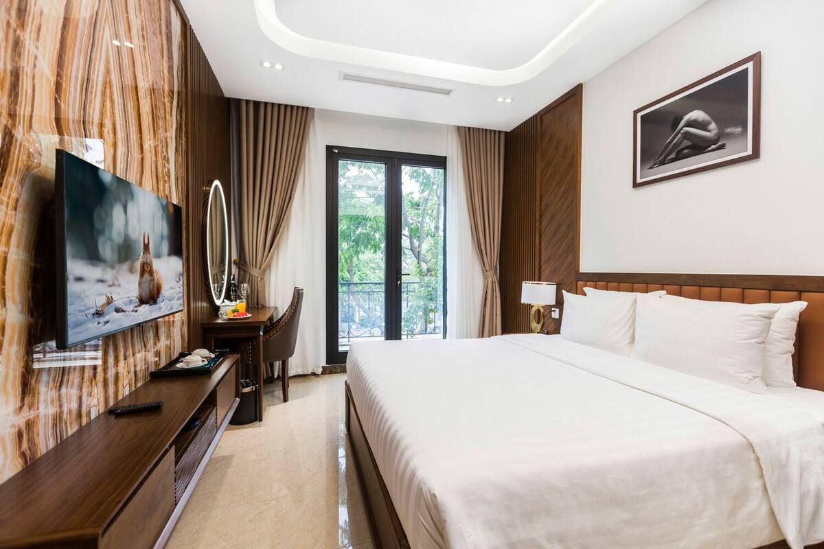 A hotel room with a large bed, a TV, a desk with a chair, a mirror, and a window with curtains.