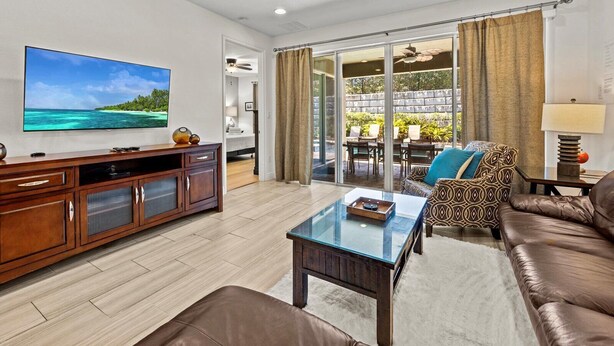 A spacious living room with a large flat-screen TV, a leather sofa, a glass coffee table, and a dining area with outdoor seating. - Luxury Renovation-Huge Upgrades! MustSee Pool -LED Games Room! Themed Rooms! (Reunion)