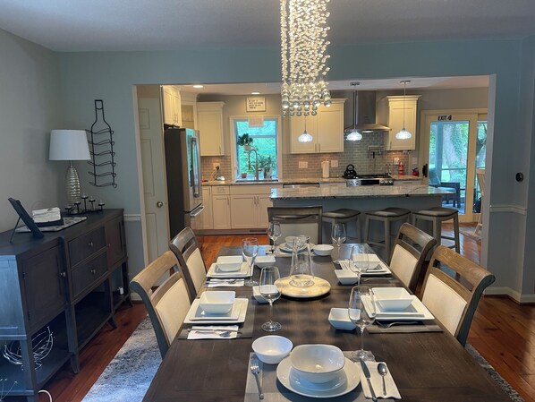 A dining area with a dark wooden table set for six, surrounded by chairs. A chandelier hangs above, and the room has a view of the kitchen and outdoor area through a glass door.