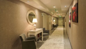 Un corridoio d'albergo dalla pianta lunga e stretta, con un’area salotto che include un tavolo, sedie, uno specchio rotondo e una lampada a parete.
