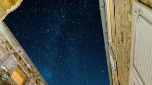 Un edificio de piedra con una escalera que lleva a un balcón, una zona de comedor con sillas y una mesa, y un cielo nocturno con estrellas visibles a través del espacio abierto.