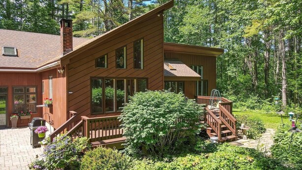 A two-story wooden cabin with a deck, surrounded by lush greenery and trees.