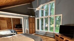 A bedroom with a large bed, wooden flooring, a ceiling fan, and a window with a view of trees.