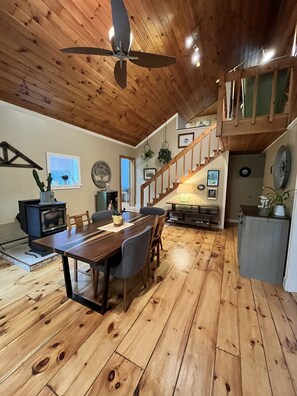 A spacious room with a wooden floor, a dining table with chairs, a ceiling fan, a staircase, and a kitchen area with a stove. - Sebago Pine Lodge +Bonus Yurt! (Windham)