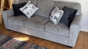 Ein graues Sofa mit gemusterten Kissen, ein gerahmtes Bild an der Wand und eine Holzleiter an der Wand.