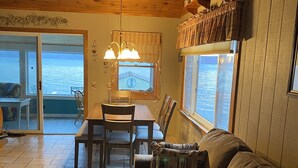 Ein gemütliches Blockhaus-Interieur mit Holzbalkendecke, einer Essecke mit Tisch und Stühlen, einer Couch mit gemusterten Kissen und Blick auf das Wasser durch die Fenster.