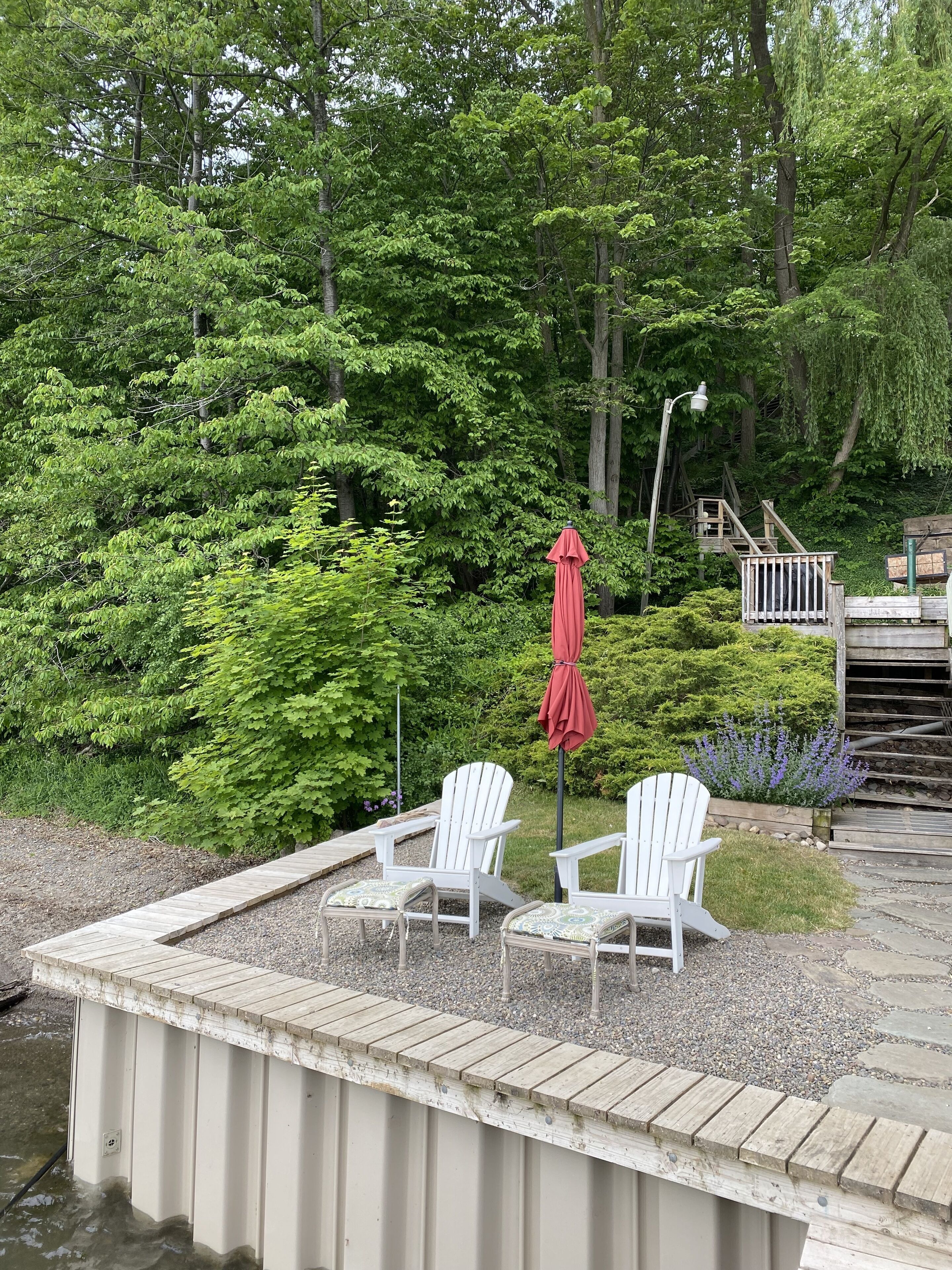 A dock with white Adirondack chairs and a red umbrella, surrounded by lush greenery and a wooden deck.