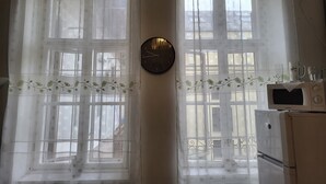 A room with two large windows covered by sheer curtains, a wall clock, and a microwave on a white cabinet. - Casa Nora Guest House (Budapest)