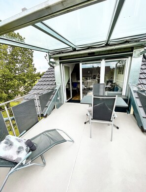 Un balcón acristalado con una mesa y sillas, vista al jardín trasero y una puerta que da a un espacio interior.