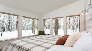 A bedroom with a large bed, multiple windows, a chair, and a view of a snowy forest. - Owl Ridge Cabins (Wausau)