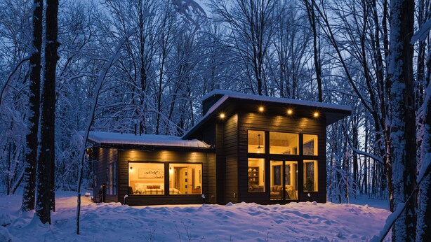 A wooden cabin with large windows and a well-lit interior, surrounded by snow-covered trees. - Owl Ridge Cabins (Wausau)