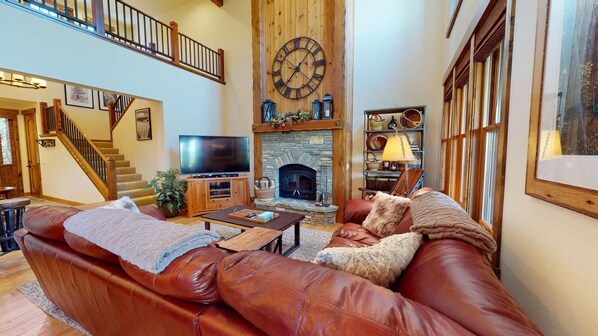 A spacious living room with a leather sofa, a stone fireplace, a large clock, and a staircase leading to an upper level.