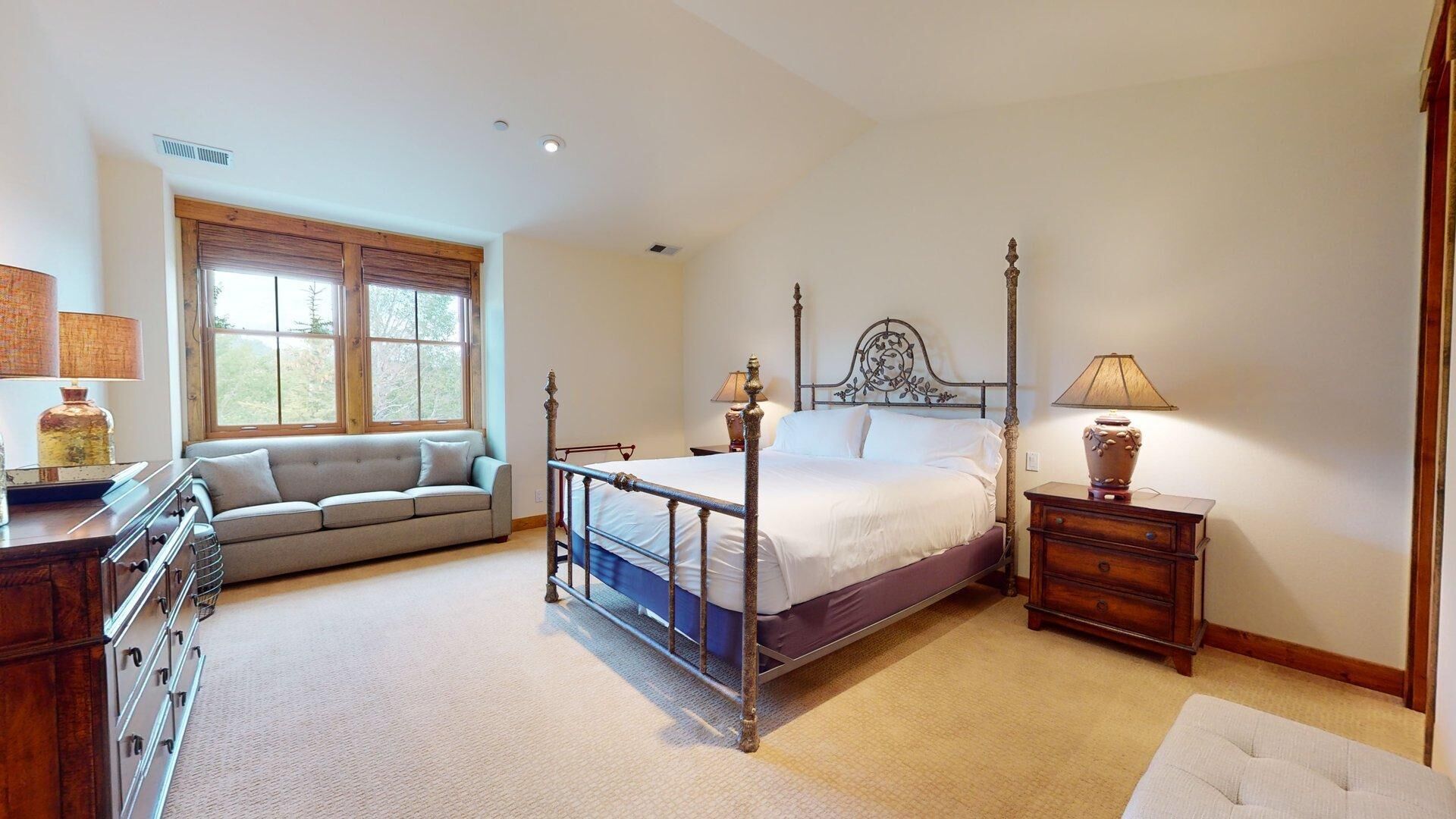 A bedroom with a four-poster bed, a sofa, a dresser, and a window with a view of trees.