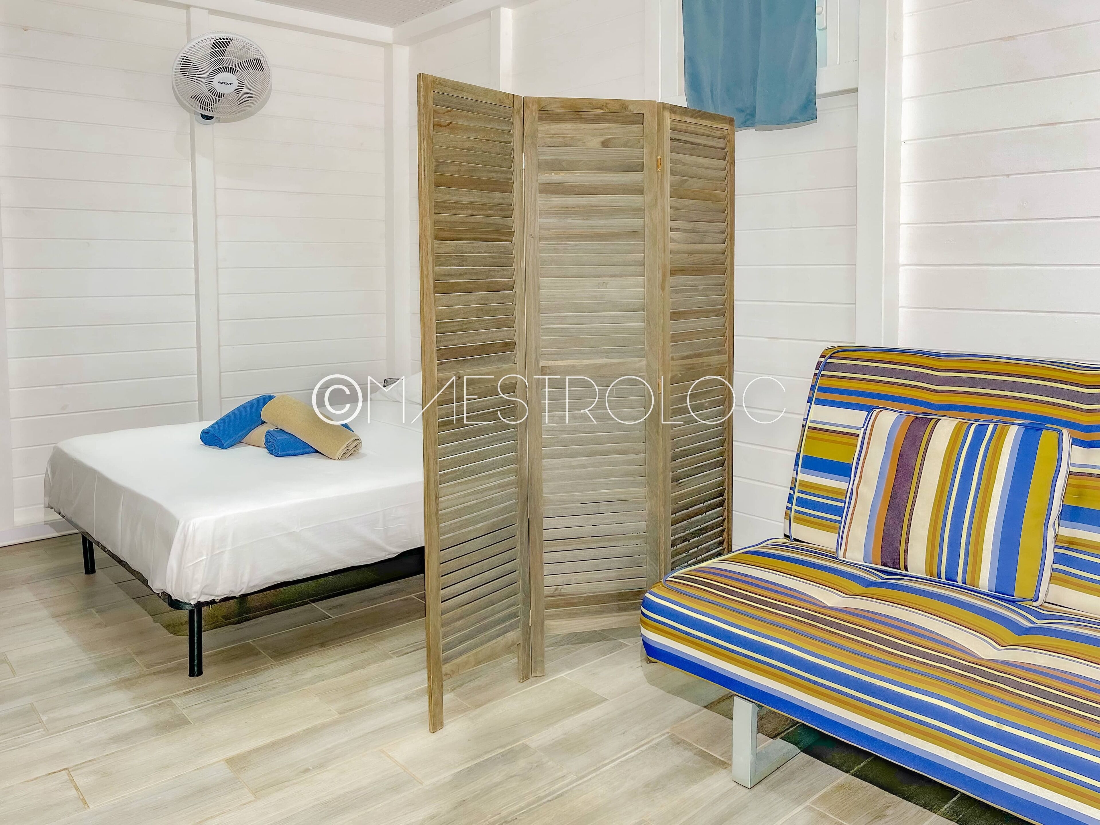 A room with a bed, a sofa, a wooden partition, and a wall-mounted fan.
