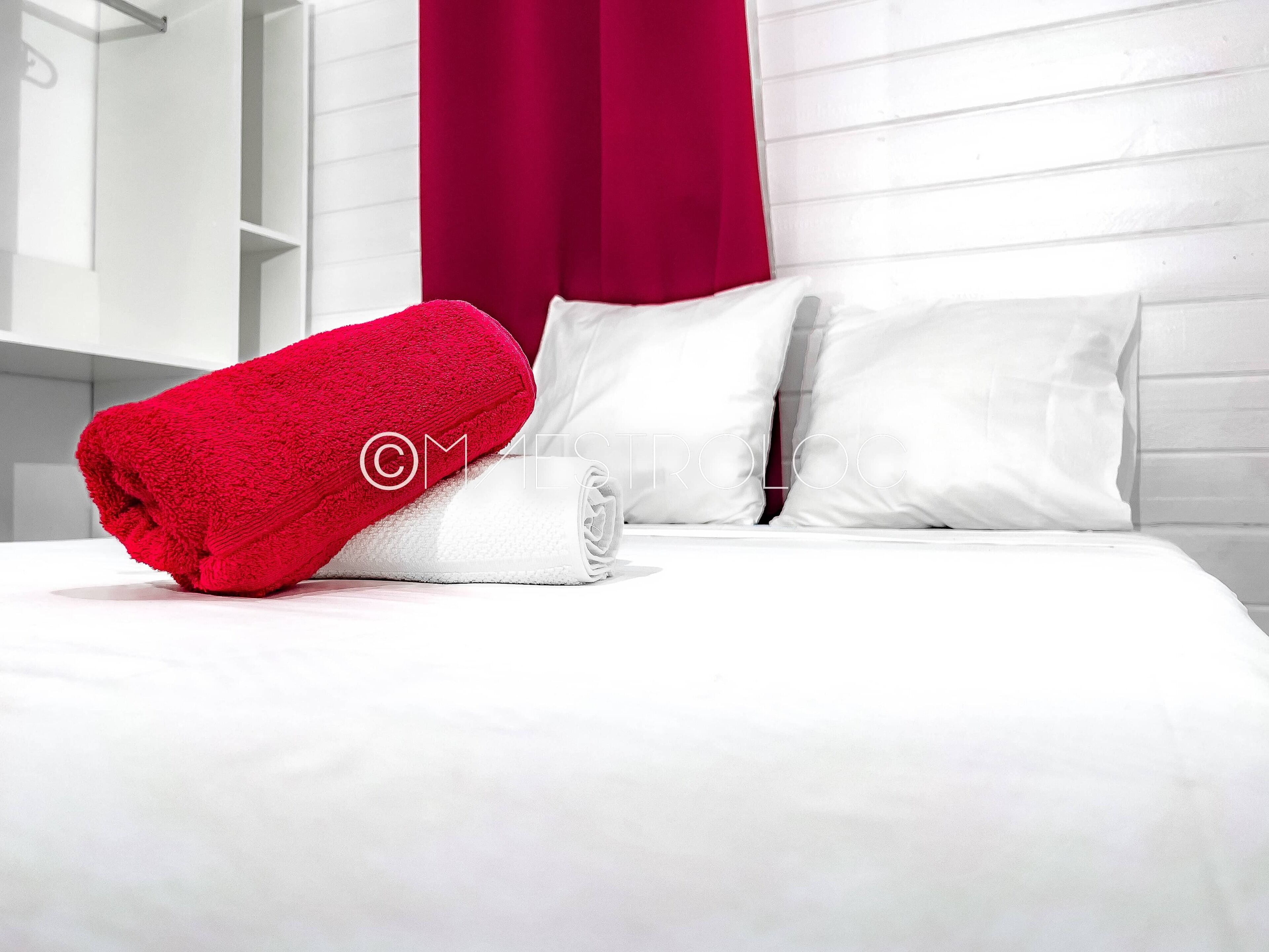 A neatly made bed with white linens and a red towel, white pillows, and a red curtain in a modern room with white walls and a wooden headboard.