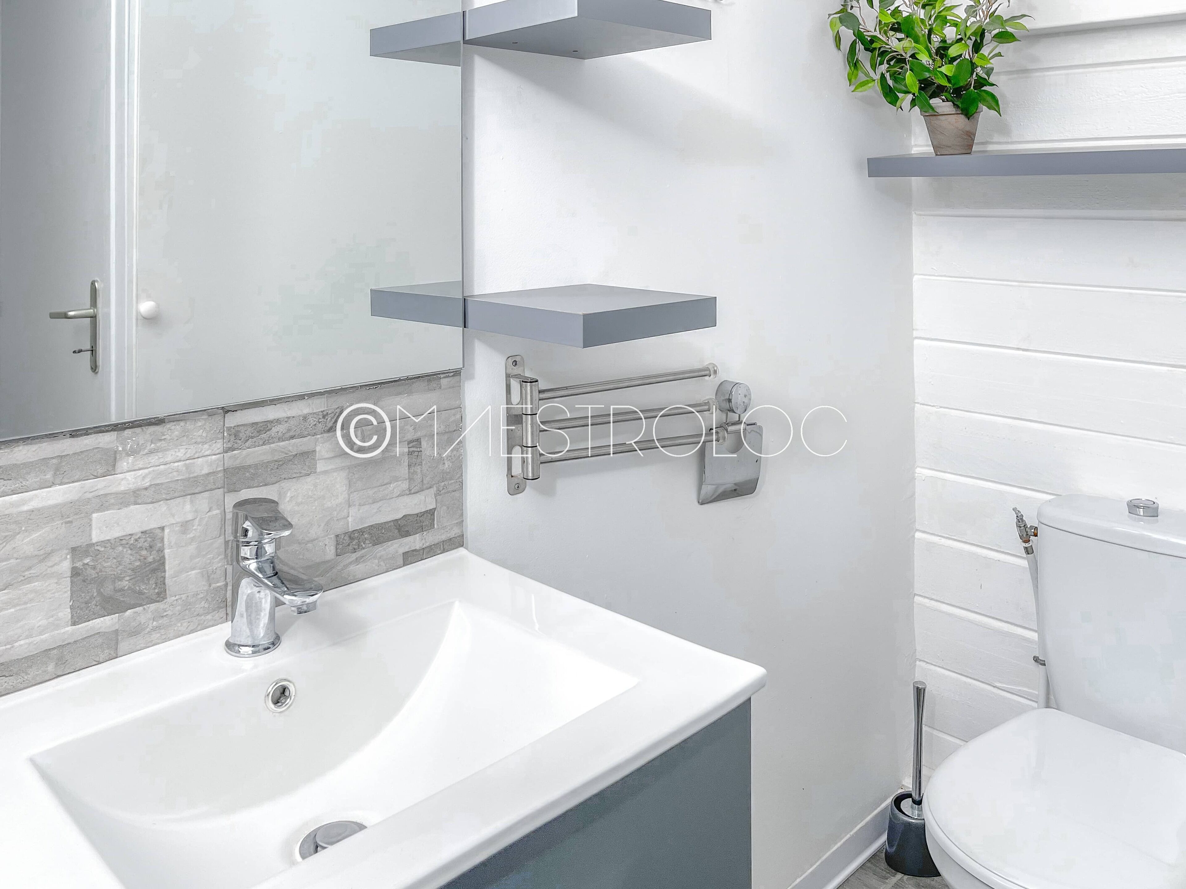 A modern bathroom with a white sink, a mirror, and a toilet. There are gray floating shelves above the sink and a potted plant on the right.