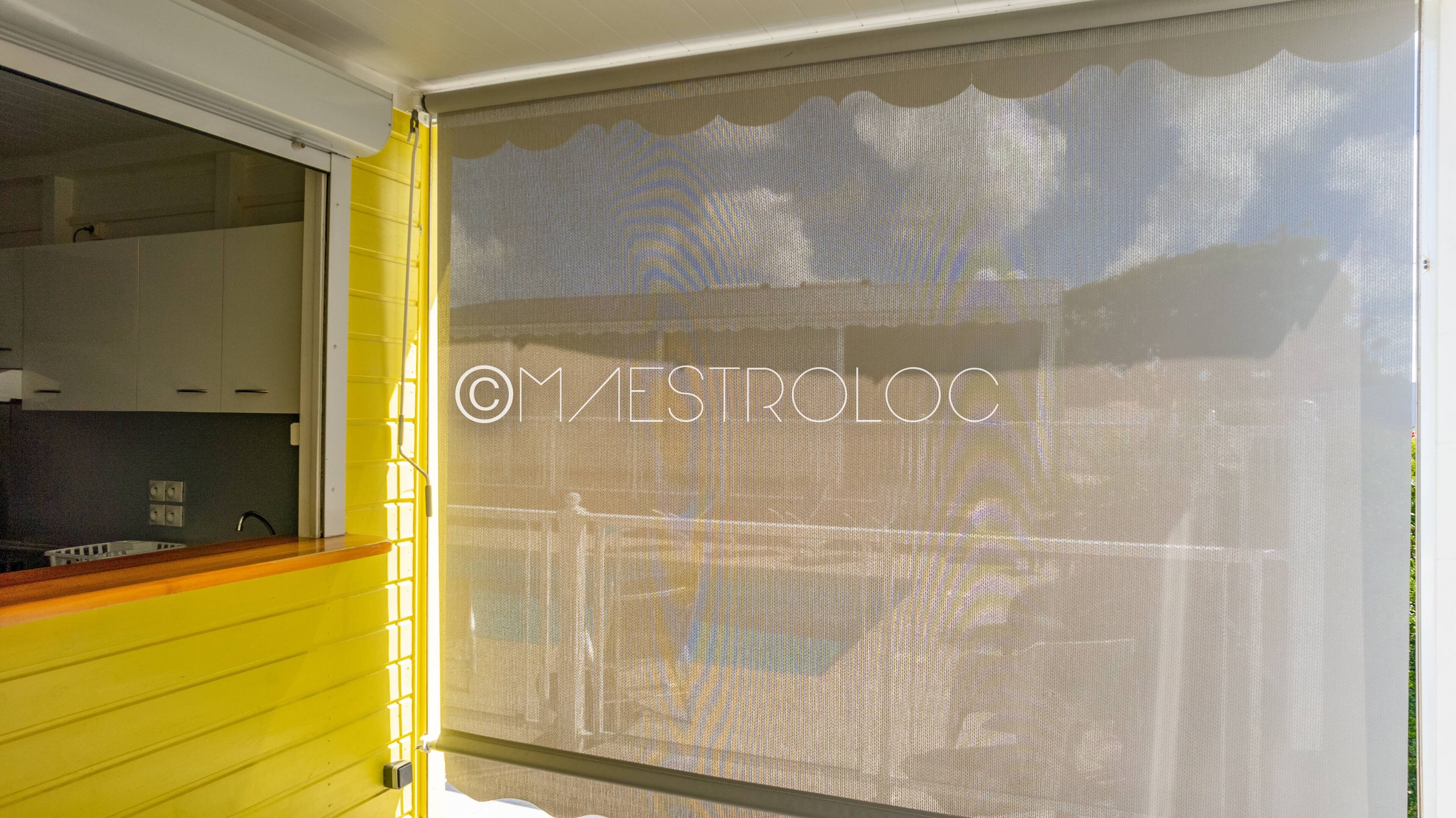 Una habitación con pared amarilla, una ventana con mosquitero y una zona de cocina visible con armarios blancos.