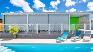 A poolside area with a covered seating area, a pool, and a building with a green door. - Gîtes Arc-en-ciel (Saint-François)