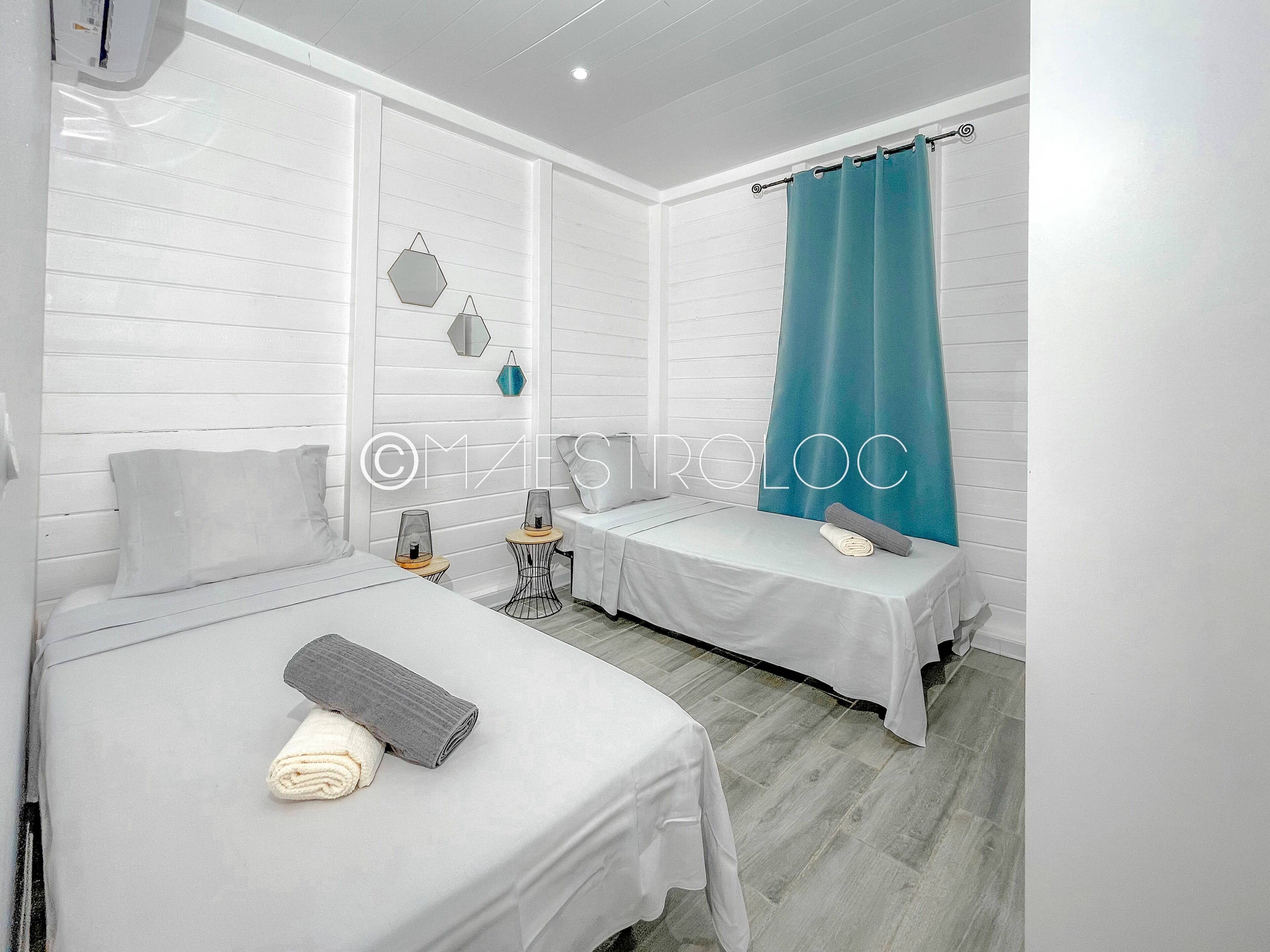 A hotel room with two beds, a large window with white shutters, a teal curtain, and a wall-mounted mirror.