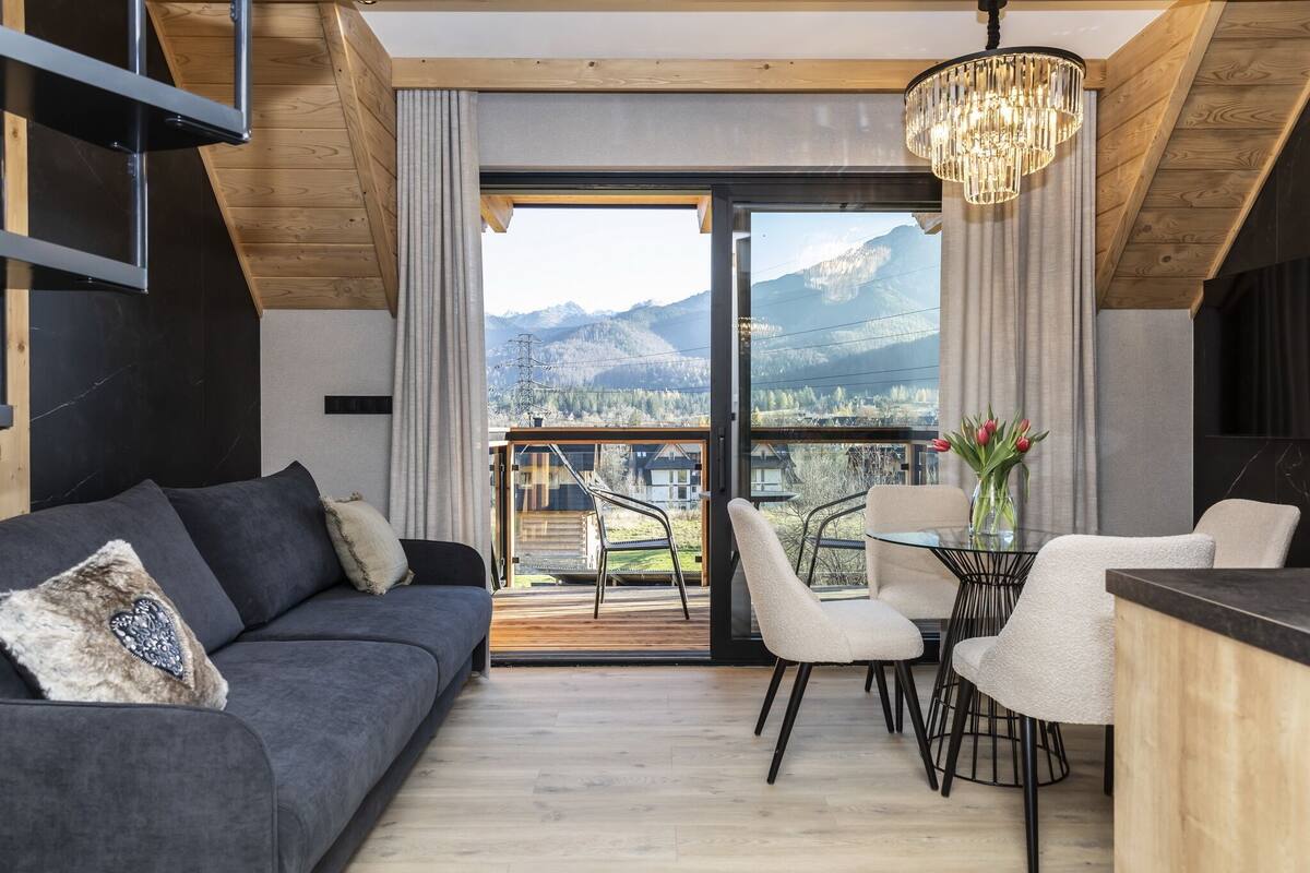 Una sala de estar moderna con un sofá gris, una mesa de comedor con sillas y vistas a montañas a través de puertas de vidrio grandes.