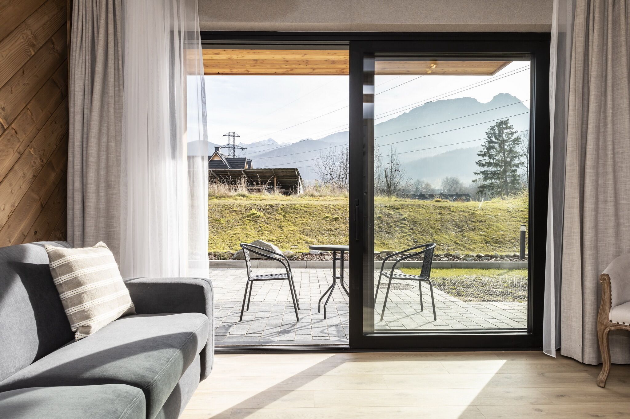 Una sala de estar moderna con un sofá y una mesa, una puerta corrediza de vidrio que da a un patio exterior con sillas y vistas a montañas.