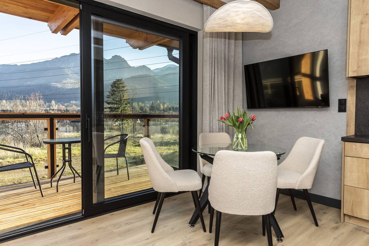 Una habitación moderna con un amplio ventanal corredero que da a una terraza exterior, un televisor de pantalla plana montado en la pared y una mesa de comedor con sillas.