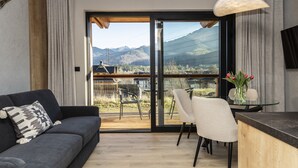 A modern living room with a sofa, a glass table, and chairs, a wooden deck, and a view of mountains.