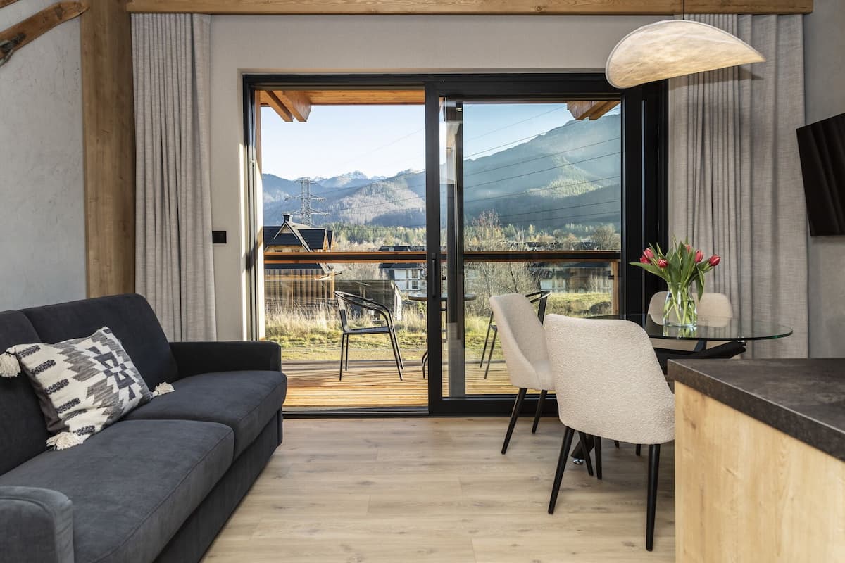 Una sala moderna con sofá, una mesa de vidrio y sillas, una terraza de madera y vistas a las montañas.