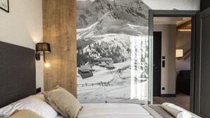 A hotel room with a large wall mural of a snowy mountain landscape, a bed with white linens, a wooden headboard, and a side table with a rolled towel.