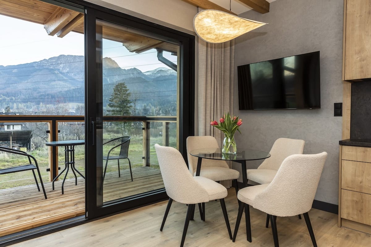 Una habitación moderna con puerta de cristal que da a un balcón, una mesa de comedor con sillas, un jarrón con flores y un televisor de pantalla plana montado en la pared.