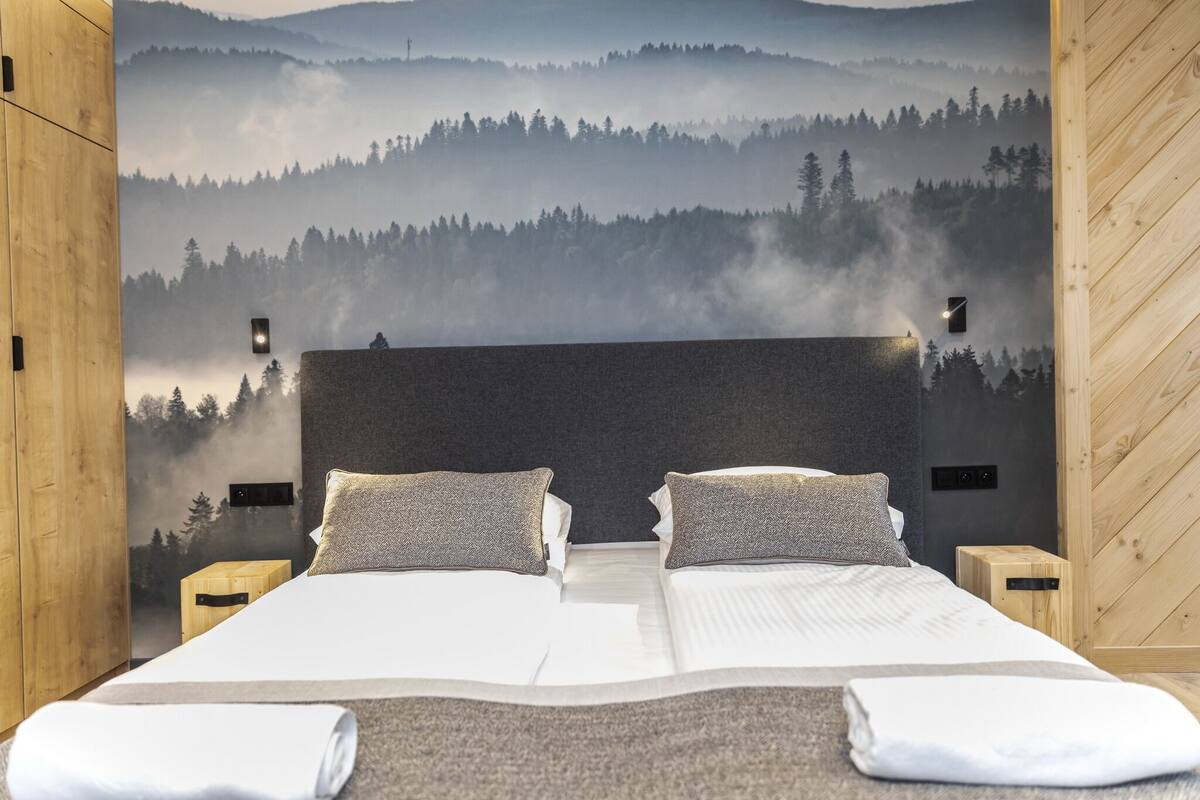 Cama doble con dos almohadas, cabecera de madera y un mural en la pared con un bosque.