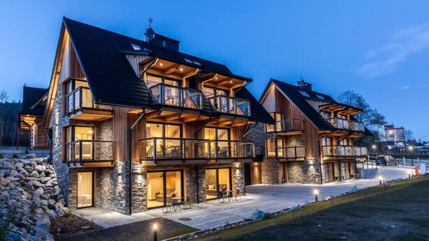 A modern multi-story house with large windows and balconies, stone exterior, and a well-lit driveway.