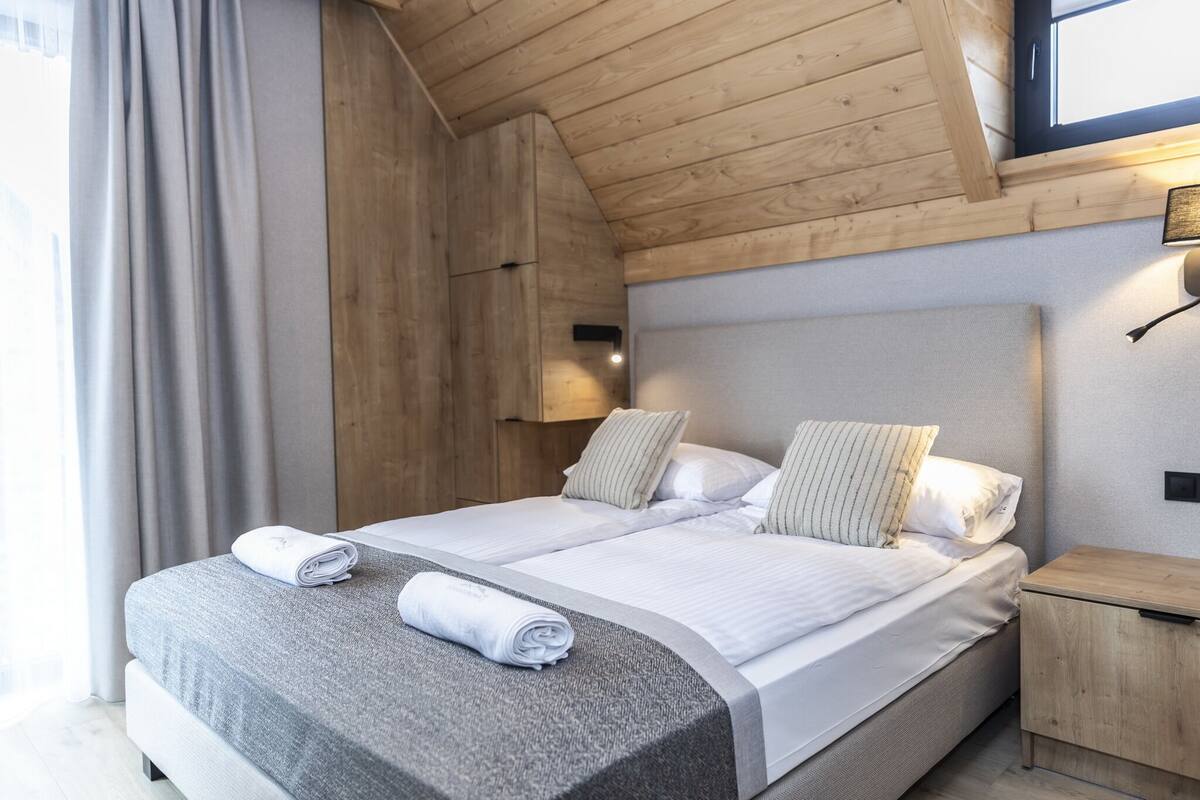 Cama doble con cabecera gris y ropa de cama blanca, mesita de noche de madera y ventana con cortinas.